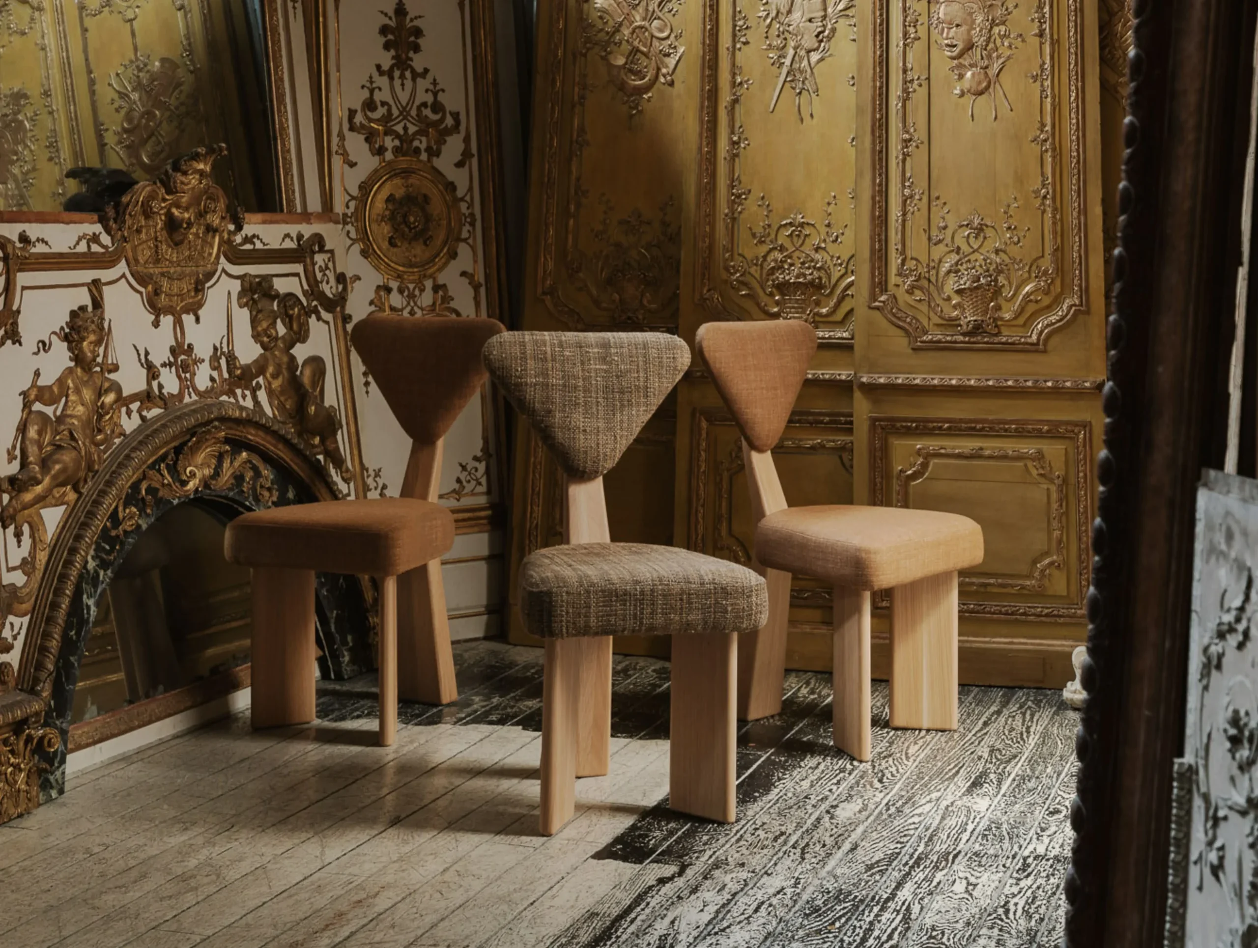 Set of three contemporary upholstered chairs with triangular backrests displayed in an ornate room with gilded wall panels and mirrors.