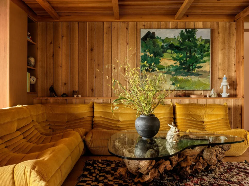 Mid-century inspired living room with mustard velvet sectional sofa, rustic wood-paneled walls, and sculptural glass coffee table.