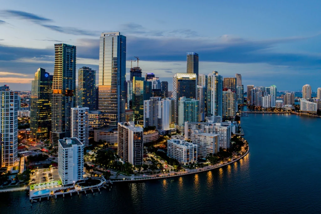Skyline view of downtown Miami with illuminated high-rise buildings along the waterfront at dusk.