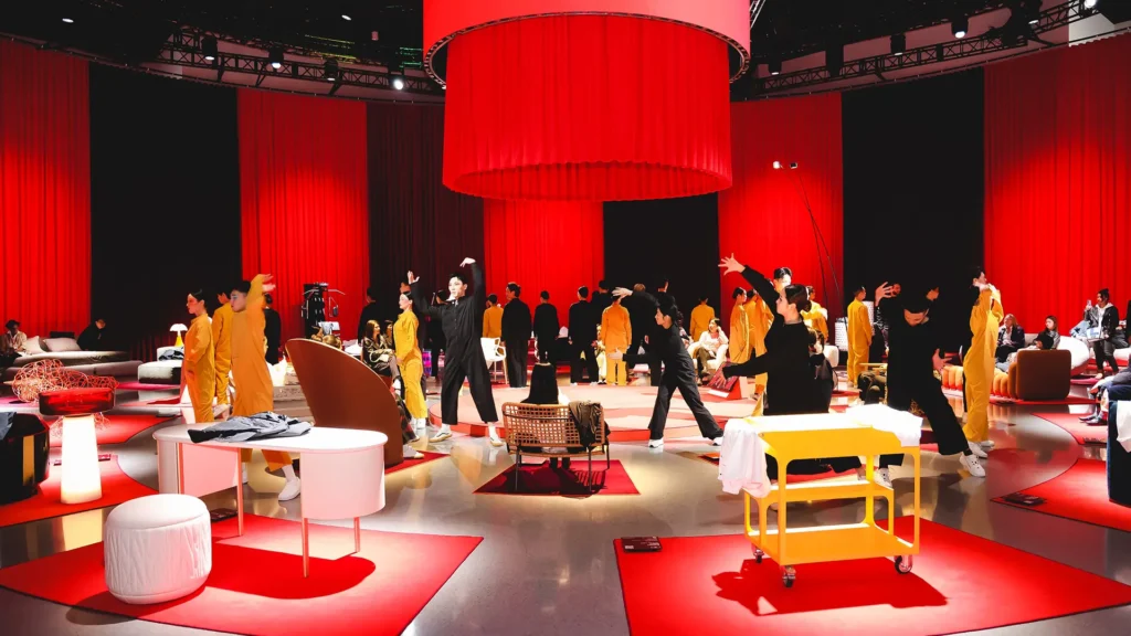 Performance art and contemporary furniture display with red backdrop and performers in black and yellow outfits.