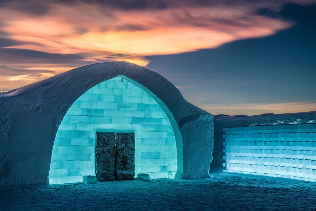 Exterior of the Icehotel in Sweden, a glowing igloo-like structure built from ice and snow, illuminated under a colorful sunset sky.