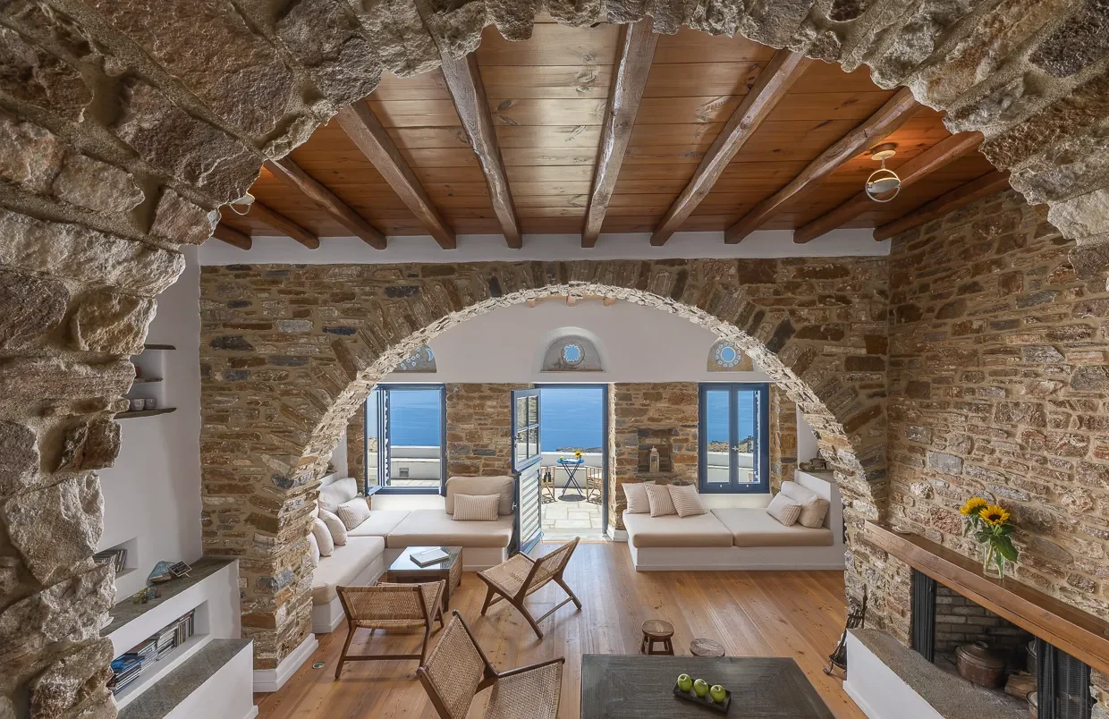 Rustic Mediterranean living room with stone arches, wood beams, and sea views through blue-framed windows