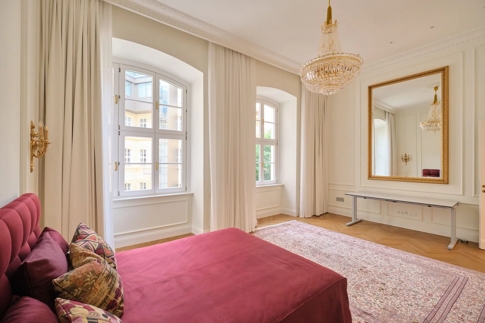 Refined bedroom in Vienna apartment with large windows, crystal chandelier, and velvet red bed.