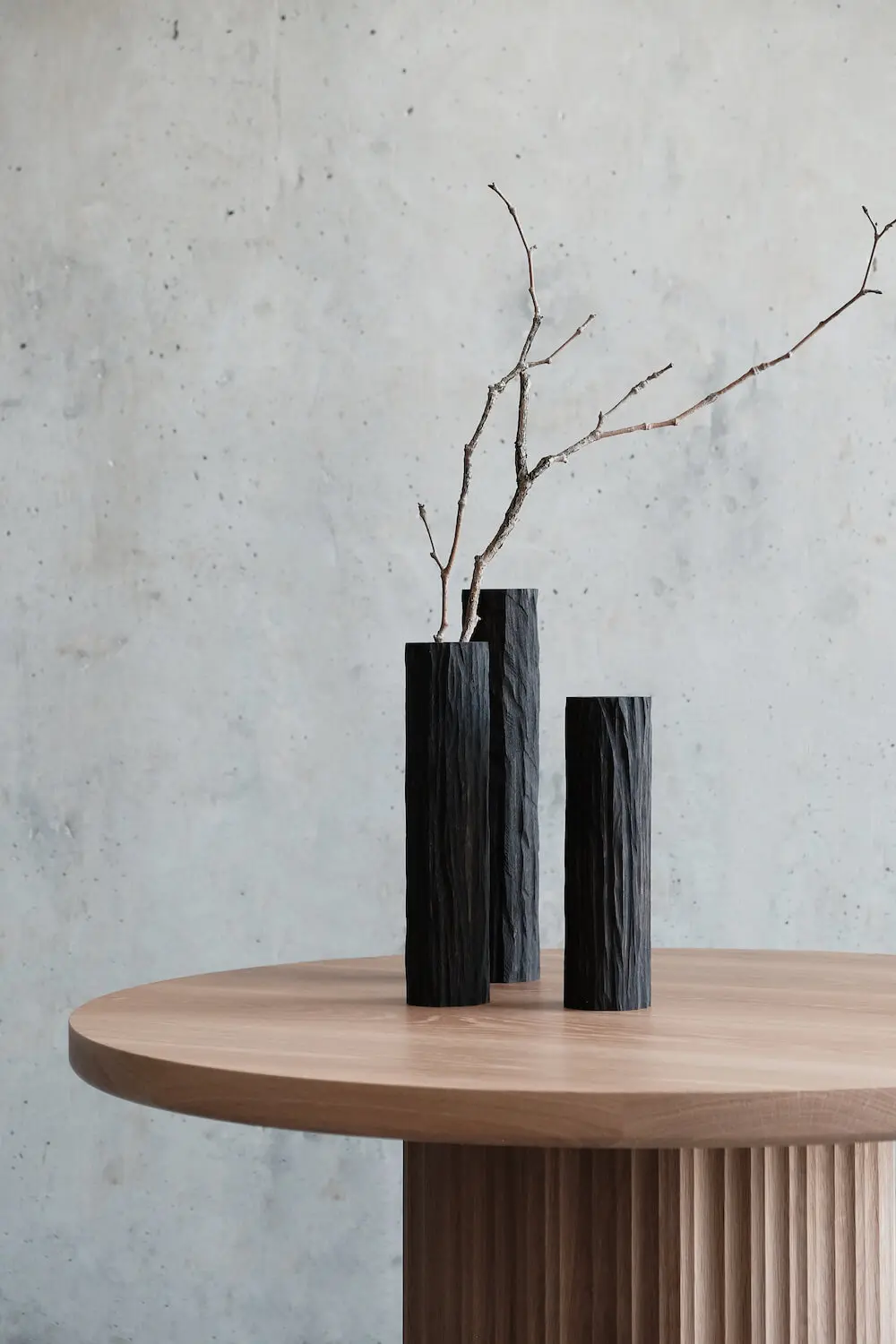 Minimalist wooden table with three charred black vases and a single dry branch against a concrete wall.