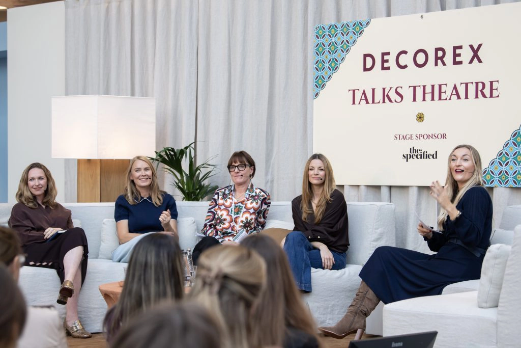 A panel of five design professionals seated on light sofas during the Decorex Talks Theatre session at Olympia London. The speakers engage in a lively discussion on contemporary interior design trends, under a banner reading “Decorex Talks Theatre – Stage Sponsor: the specified.”