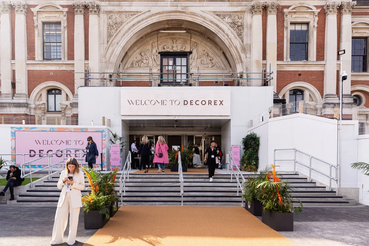 Entrance to Decorex 2023 at Olympia London, featuring the grand neoclassical façade of the venue with ornate stone carvings and columns. Visitors walk up the carpeted steps beneath the “Welcome to Decorex” sign, surrounded by lush greenery and colorful event signage.