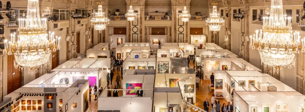 Art & Antique 2025 exhibition inside Vienna’s historic Hofburg, with illuminated gallery stands beneath grand crystal chandeliers.