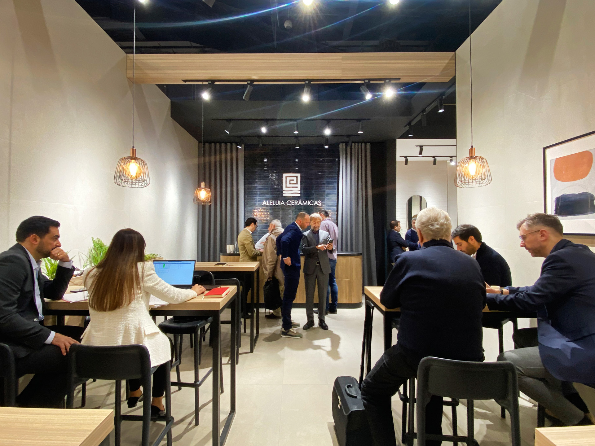 Visitors meeting inside the Aleluia Cerâmicas stand at Cevisama 2024, surrounded by ceramic wall displays and warm pendant lighting.