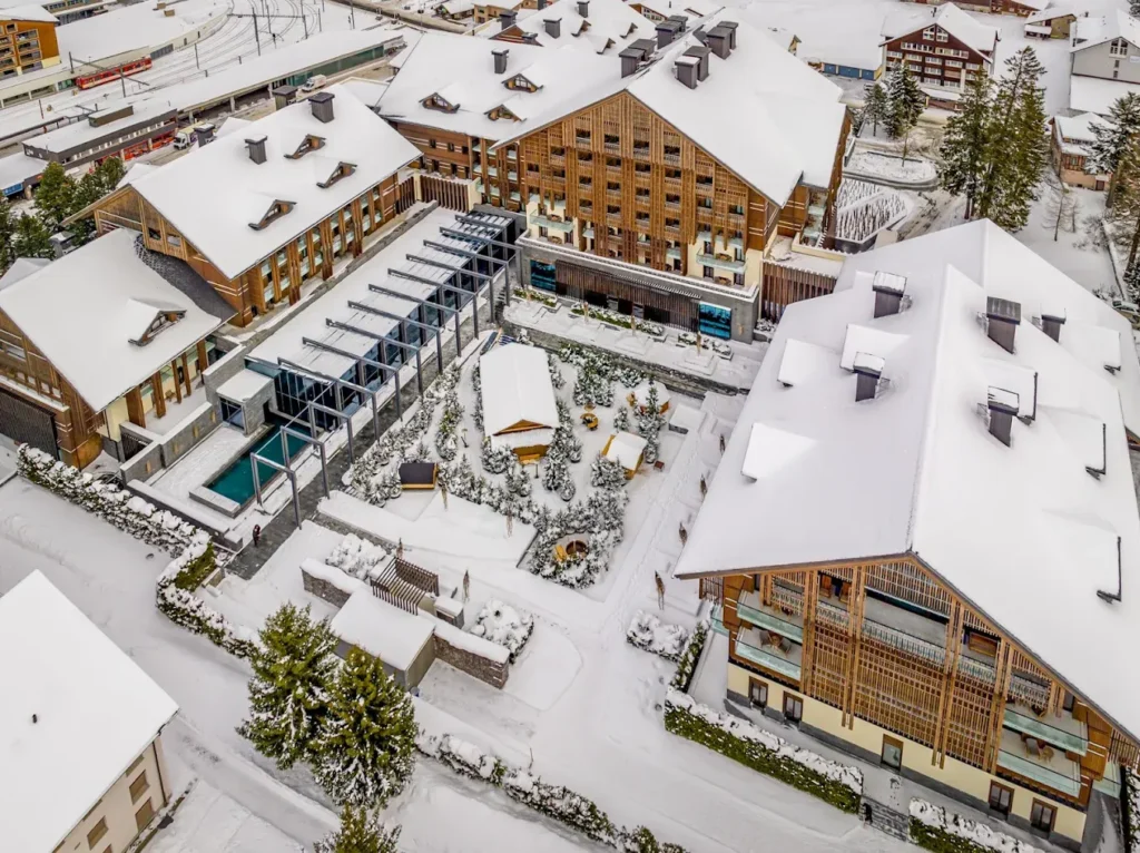 Aerial winter view of The Chedi Andermatt in Switzerland, featuring contemporary chalet-inspired architecture surrounded by snow covered roofs and mountain scenery.